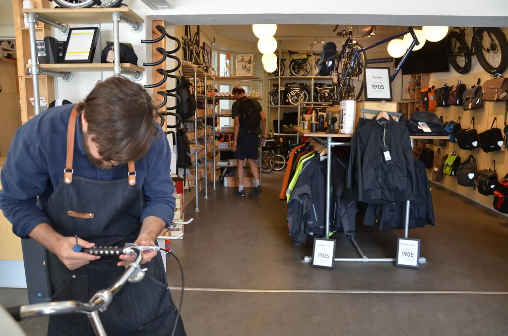 A mechanic in a work apron adjusting a bicycle's handlebars inside a bike shop, with bikes and cycling clothing on display in the background.