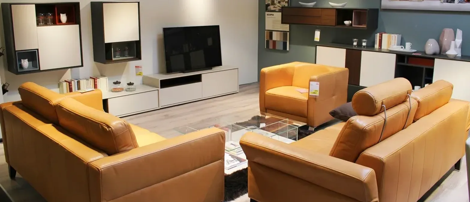 A furniture showroom display of tan leather sofas and an armchair arranged in front of low wall cabinets and a wall-mounted television.