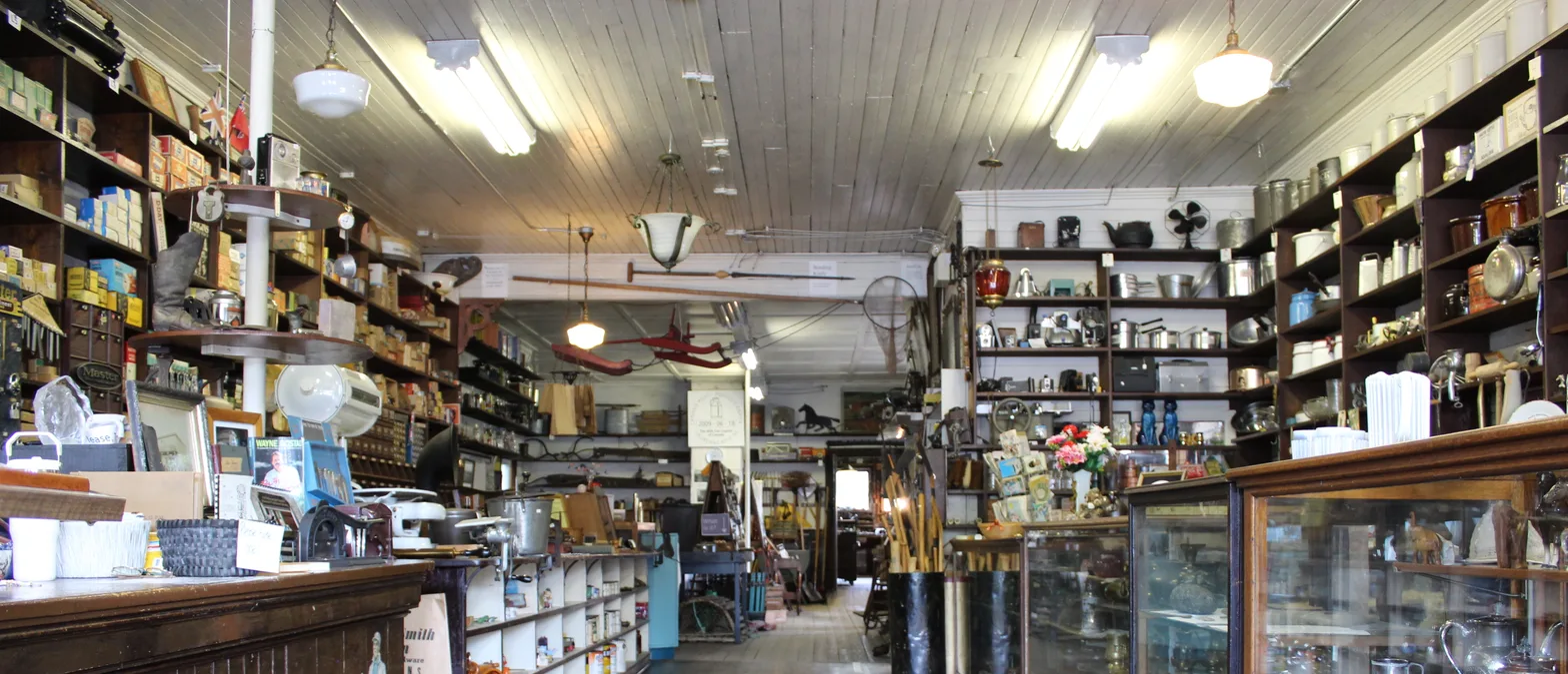 The inside of an old general and hardware store, with long wooden counters and glass display cases in front of shelves stacked with boxes, kitchenware and tools.