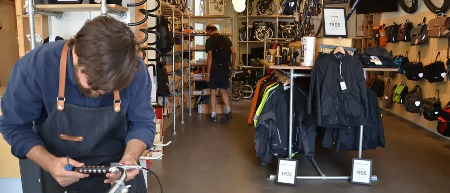 A shop employee in a canvas apron adjusts a bicycle's handlebar controls at a repair bench inside a small shop.