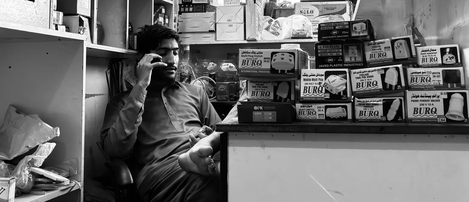A shopkeeper in a small hardware shop holding a phone to his ear, seated behind a counter stacked with boxed plugs and sockets.